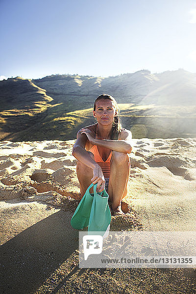 Porträt einer selbstbewussten Frau  die am Strand sitzend Tauchflossen hält