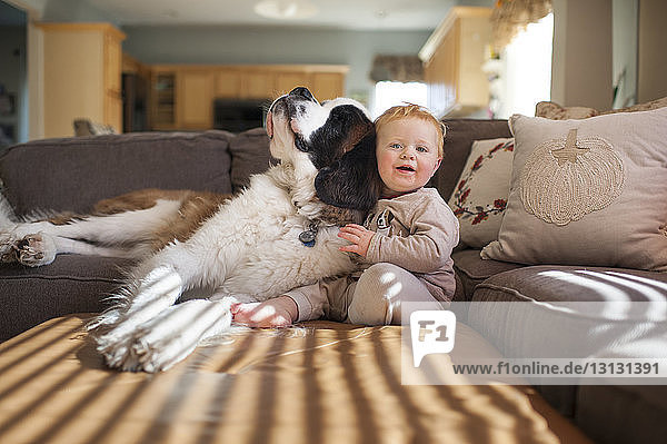 Porträt eines süßen kleinen Jungen  der mit seinem Hund auf der Couch im heimischen Wohnzimmer spielt