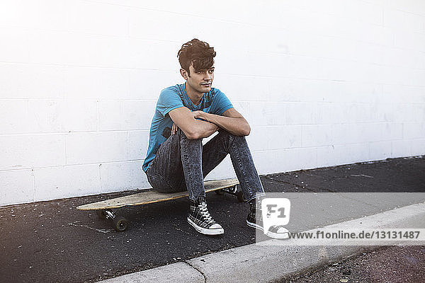 Nachdenklicher Mann sitzt auf Skateboard auf dem Bürgersteig vor weißer Wand