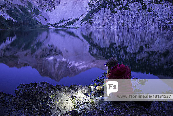 High angle view of female hiker using headlamp while making coffee by lake against mountains