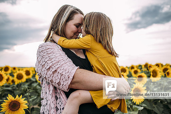 Mutter umarmt Tochter  während sie in Sonnenblumenfarm gegen den Himmel steht