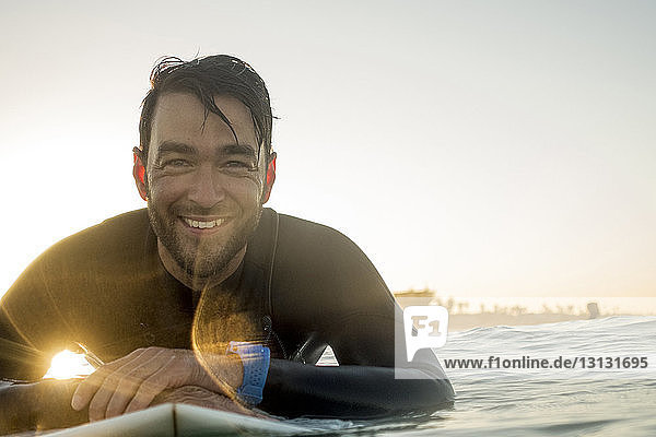 Porträt eines glücklichen männlichen Surfers  der bei Sonnenuntergang auf einem Surfbrett im Meer liegt