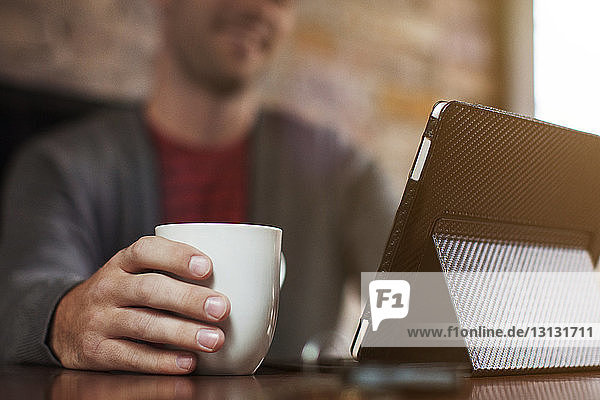 Nahaufnahme eines Mannes mit digitalem Tablett beim Kaffeetrinken am Tisch