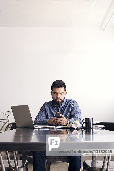 Serious man using phone while sitting at table against wall in office