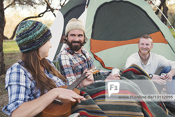 Glückliche Männer sehen eine Frau an  die vor einem Zelt im Wald Gitarre spielt