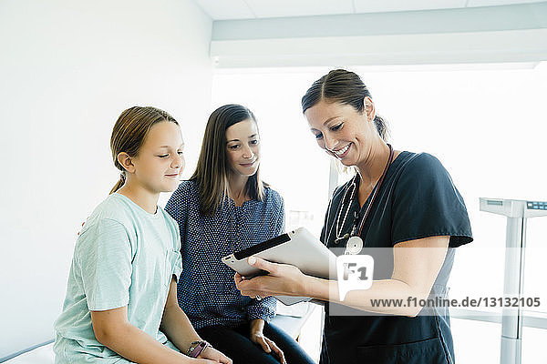 Fröhliche Kinderärztin zeigt Mutter und Tochter  die im Krankenhaus auf dem Untersuchungstisch sitzen  einen Tablet-Computer