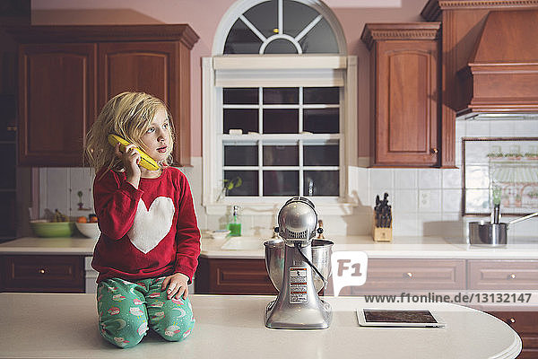 Cute girl playing with banana at table by appliance in kitchen