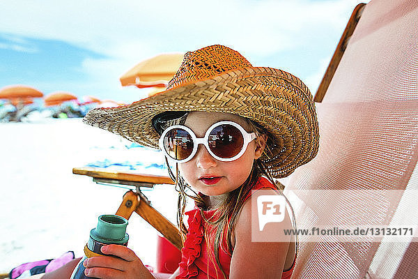 Bildnis eines Mädchens mit Sonnenbrille und Strohhut auf einem Liegestuhl am Strand sitzend