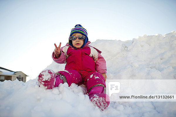 Girl in sunglasses showing peace sign while sitting on snow against clear sky