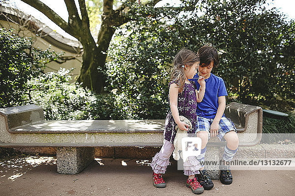 Schwester hält Stofftier  während der Bruder auf einer Parkbank sitzt