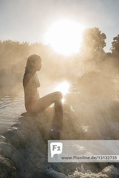 Seitenansicht einer Frau  die bei Sonnenschein auf Felsen in einem dampfabgebenden Thermalbad sitzt