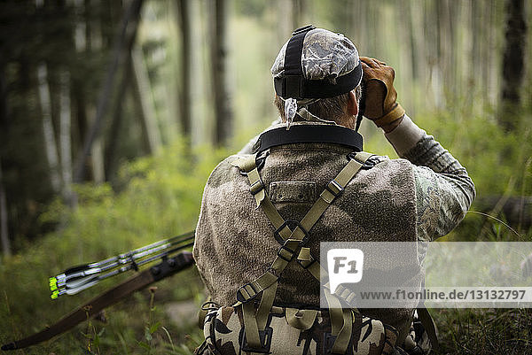 Rückansicht eines Jägers  der bei der Jagd im Wald durch ein Fernglas blickt