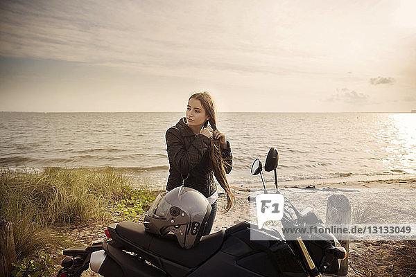 Junge Frau bindet sich die Haare  während sie mit dem Motorrad am Strand gegen das Meer steht