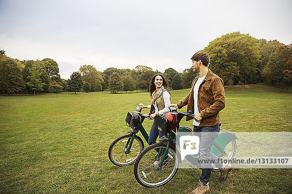 Happy couple talking while walking with bicycles on grassy field