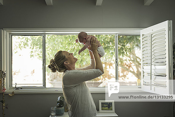 Mother playing with newborn daughter while lifting her at home against window