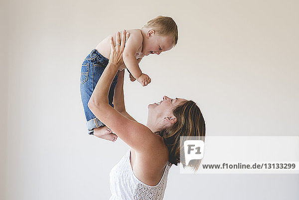 Side view of cheerful mother lifting shirtless son against white background