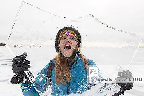 Porträt einer verspielten Frau mit herausgestreckter Zunge  die im Winter durch die Eisdecke gesehen wird