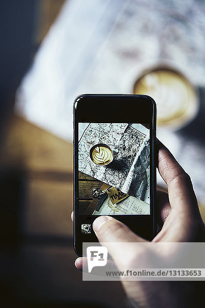 Ausgeschnittene Hand eines Mannes fotografiert Kaffeetasse mit Karte auf Tisch mit Mobiltelefon zu Hause