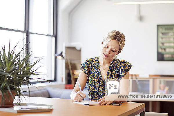 Businesswoman writing on notepad while sitting at table in office