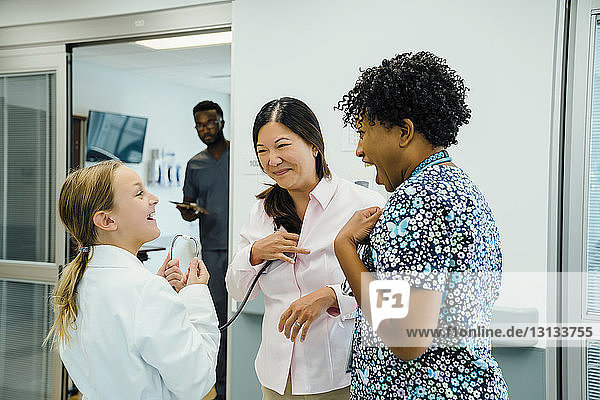 Playful girl looking at female doctor while using stethoscope on pediatrician in hospital corridor