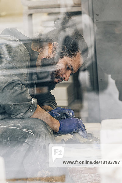 Craftsman using pottery wheel while making earthenware in workshop seen through window