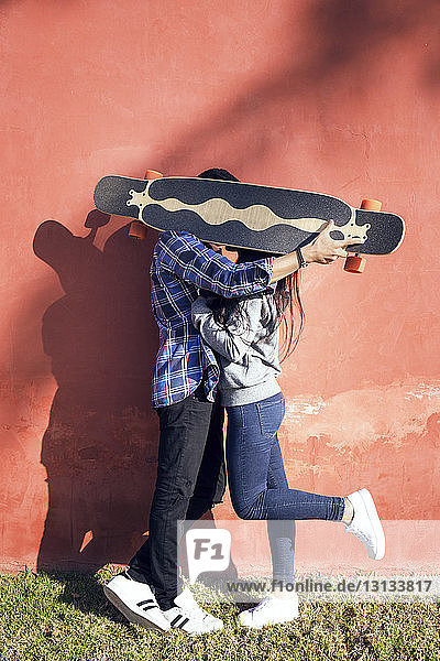 Liebespaar hält Skateboard vor dem Gesicht  während es an roter Wand steht