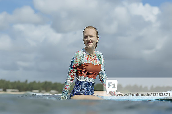 Lächelnde Frau schaut weg  während sie auf einem Surfbrett im Meer vor bewölktem Himmel sitzt