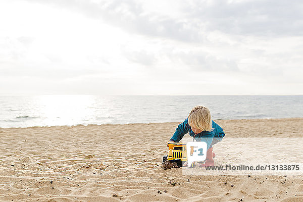 Junge spielt mit Spielzeuglastwagen am Strand gegen bewölkten Himmel