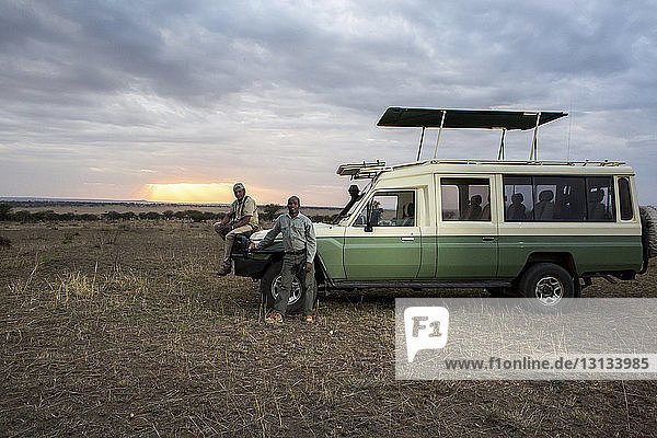 Porträt von Freunden im Geländewagen vor bewölktem Himmel im Serengeti-Nationalpark bei Sonnenuntergang