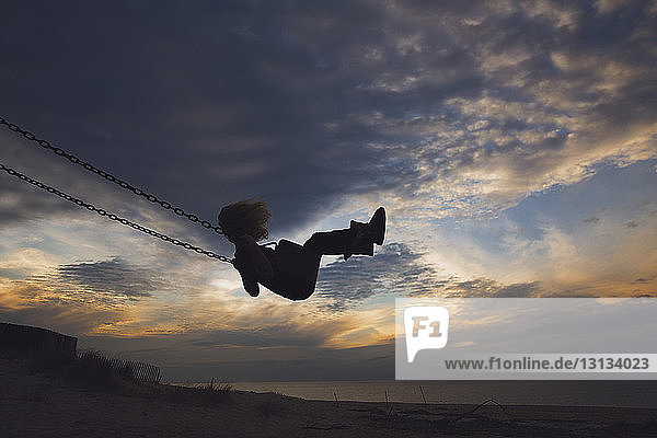 Side view of girl playing on swing at beach against cloudy sky during sunset