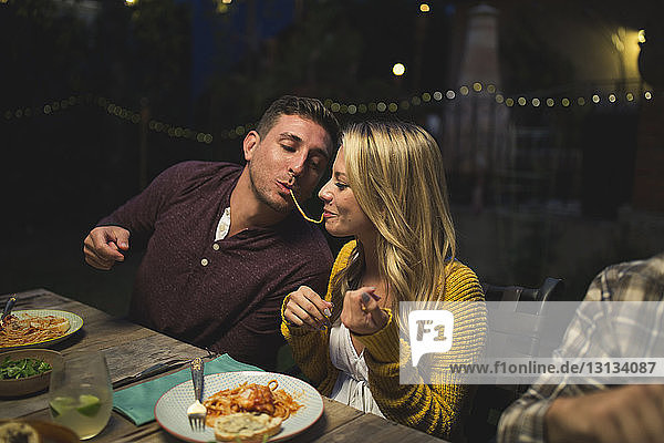 Verspieltes Paar isst Pasta und genießt gleichzeitig eine Dinnerparty mit Freunden