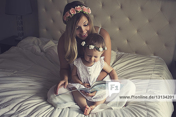 Happy mother reading story book for daughter while sitting on bed at home