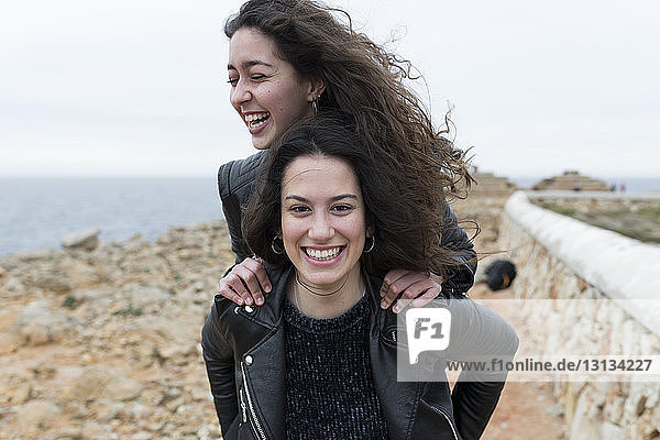 Portrait of happy woman piggybacking friend at beach against sky