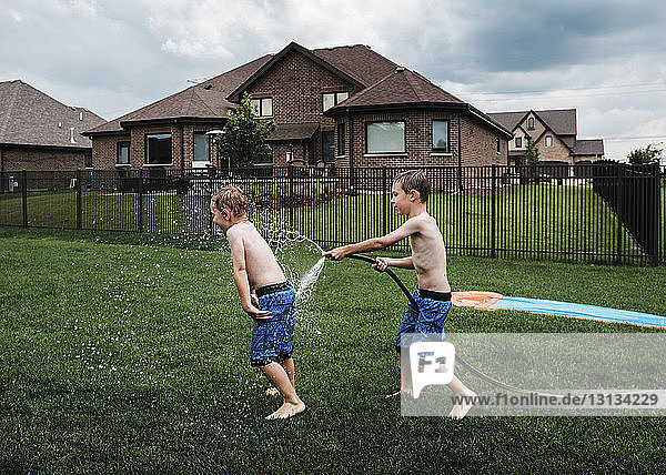 Seitenansicht eines Jungen ohne Hemd  der im Park mit einem Gartenschlauch Wasser auf seinen Bruder spritzt