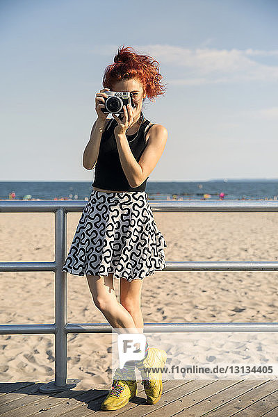 Junge Frau fotografiert durch eine Kamera  während sie am Strand am Geländer steht