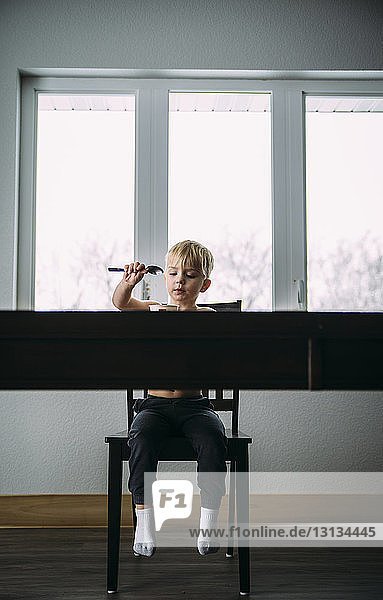 Shirtless boy eating while sitting on chair by window at home