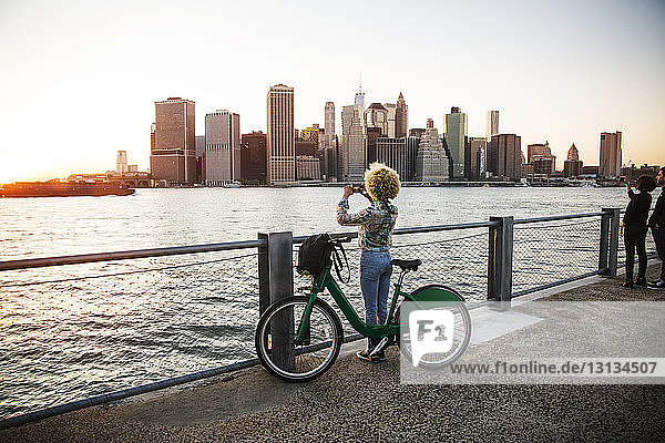 Woman standing by bicycle photographing river and city