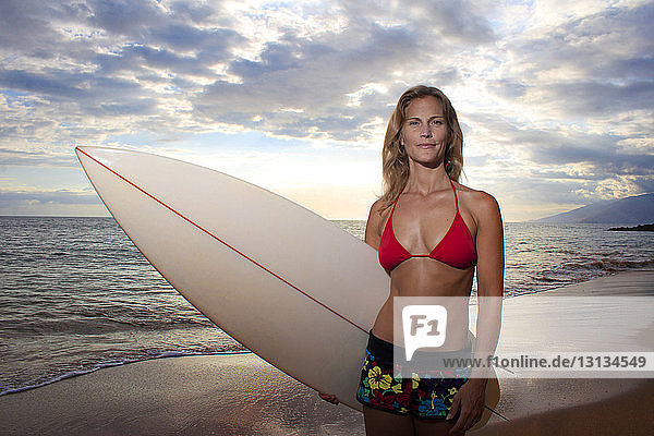 Zuversichtliche Frau mit Surfbrett am Meeresufer vor bewölktem Himmel