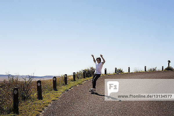 Junger Mann mit erhobenen Armen beim Skateboarden auf der Straße gegen den klaren Himmel