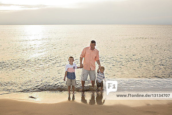 Father holding sons while standing on shore at beach during sunset