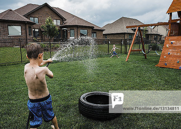 Rückansicht eines Jungen ohne Hemd  der mit einem Gartenschlauch Wasser auf seinen Bruder im Park spritzt