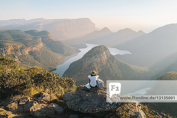 Rear view of woman looking at view while sitting on mountain against sky during sunset