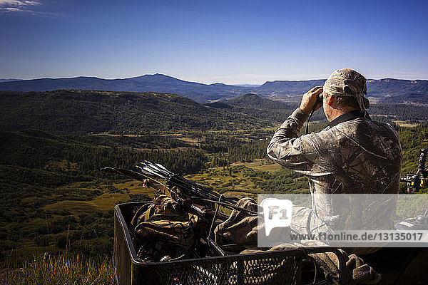 Hunter using binoculars while sitting on quadbike against sky