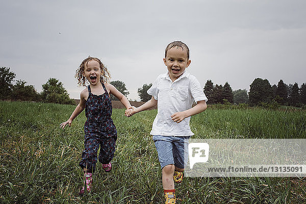 Portrait of happy wet siblings holding hands while running on grassy field against sky at park during rainy season
