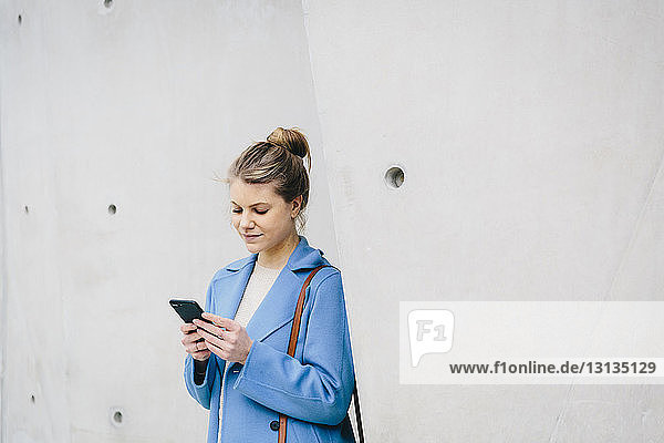 Junge Frau benutzt Smartphone  während sie in der Stadt an der Wand steht