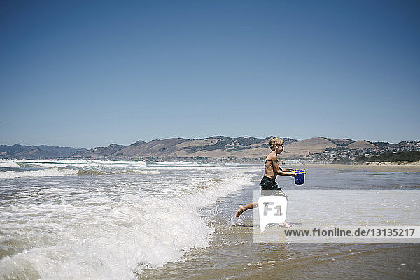 Side view of boy carrying water in bucket while running on shore at beach
