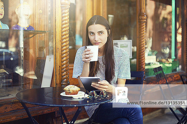 Frau trinkt Kaffee  während sie ein digitales Tablet in der Hand hält und im Straßencafé sitzt