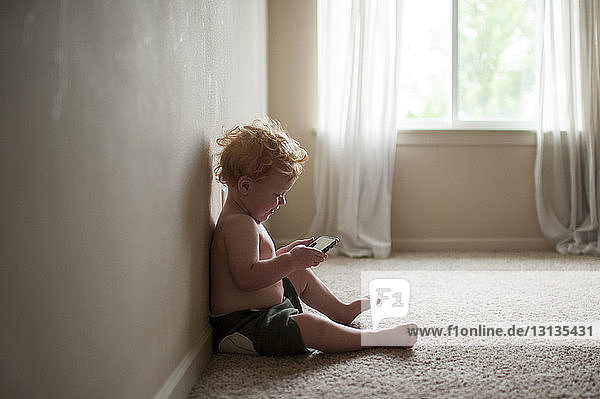 Seitenansicht eines hemdlosen  süßen Jungen  der ein Smartphone benutzt  während er zu Hause auf dem Teppich sitzt