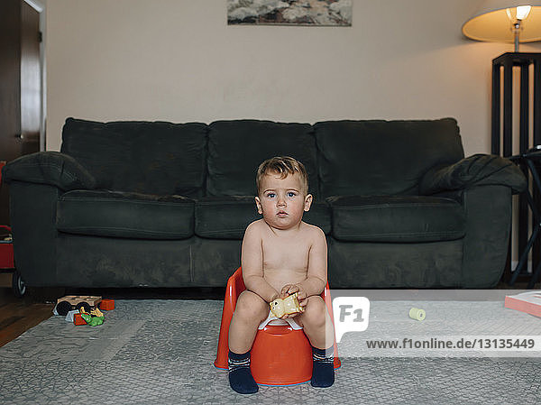 Porträt eines niedlichen Jungen ohne Hemd  der auf dem Töpfchen sitzend gegen das Sofa im Wohnzimmer uriniert