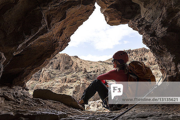 Cheerful female backpacker sitting on entrance of cave in rock formation
